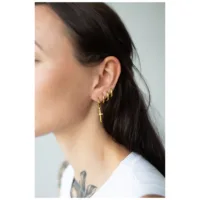 Kultaiset Little Sword Hoops -korvakorut Nainen käyttää kultaisia Little Sword Hoops -korvakoruja klipsikiinnityksellä vaalealla taustalla