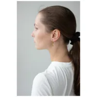 Nainen ruskealla poninhännällä käyttää kultaisia kolmionmuotoisia korvakoruja harmaata taustaa vasten