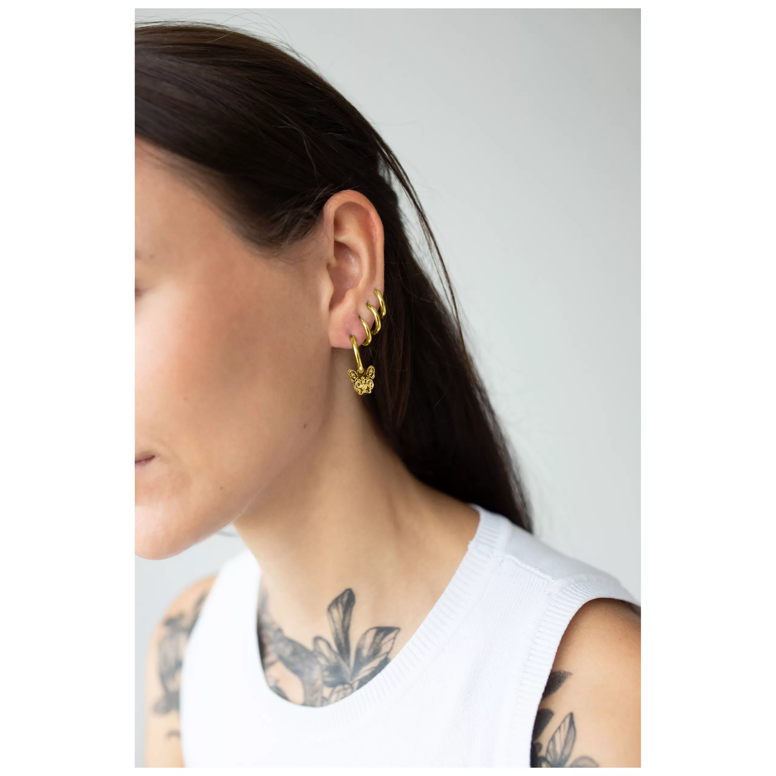 Nainen kultaisilla Little Bulldog Hoops -korvakoruilla ja tatuoinneilla valkoisella taustalla