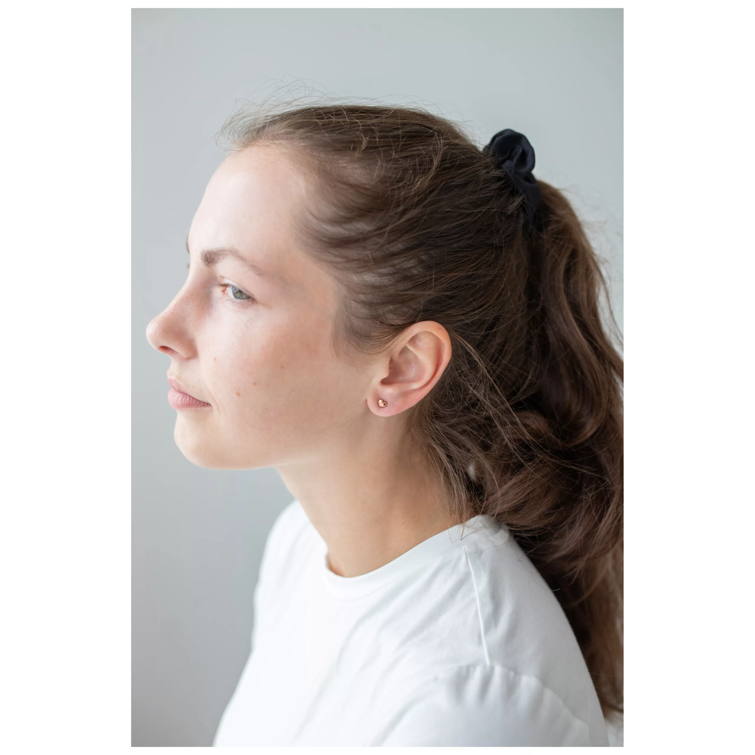 Nainen ruusukultaisilla sydänkorvakoruilla, hiukset poninhännällä, valkoinen paita