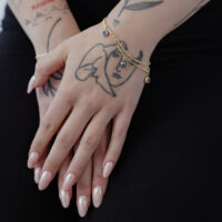 Little Nazar Armband in Gold auf Hand Goldenes Little Nazar Armband um ein Handgelenk mit Tattoos auf schwarzem Hintergrund