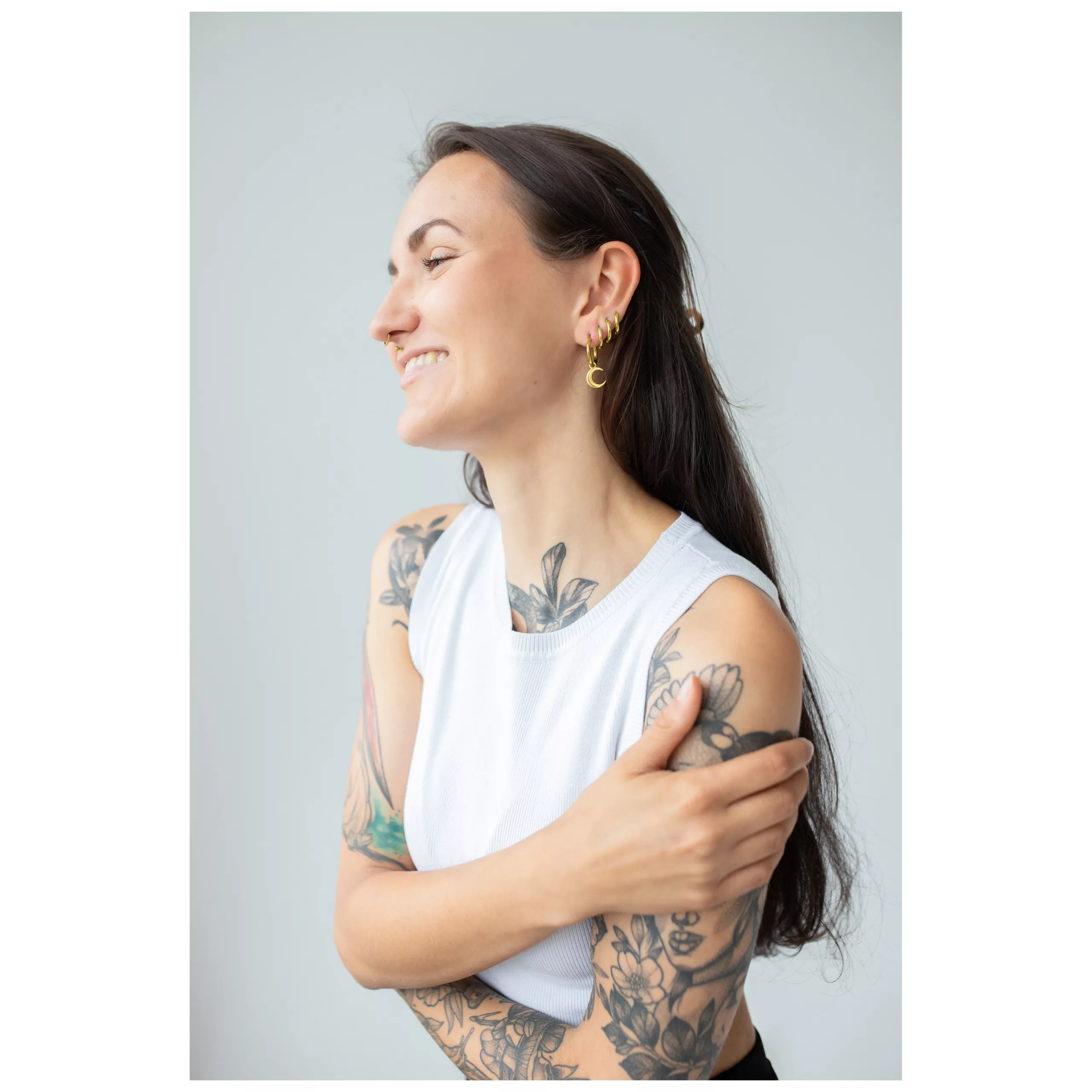 Nainen kultaisilla Little Moonchild Hoops -korvakoruilla ja tatuoinneilla, hymyilevä