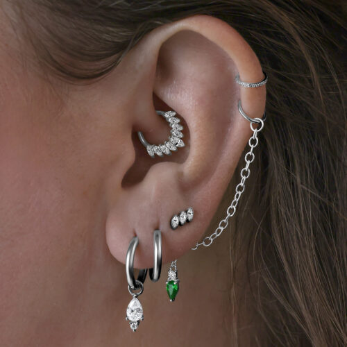Useita korvakoruja korvassa, mukaan lukien mustat Little Tear Hoops vihreällä kristallilla.