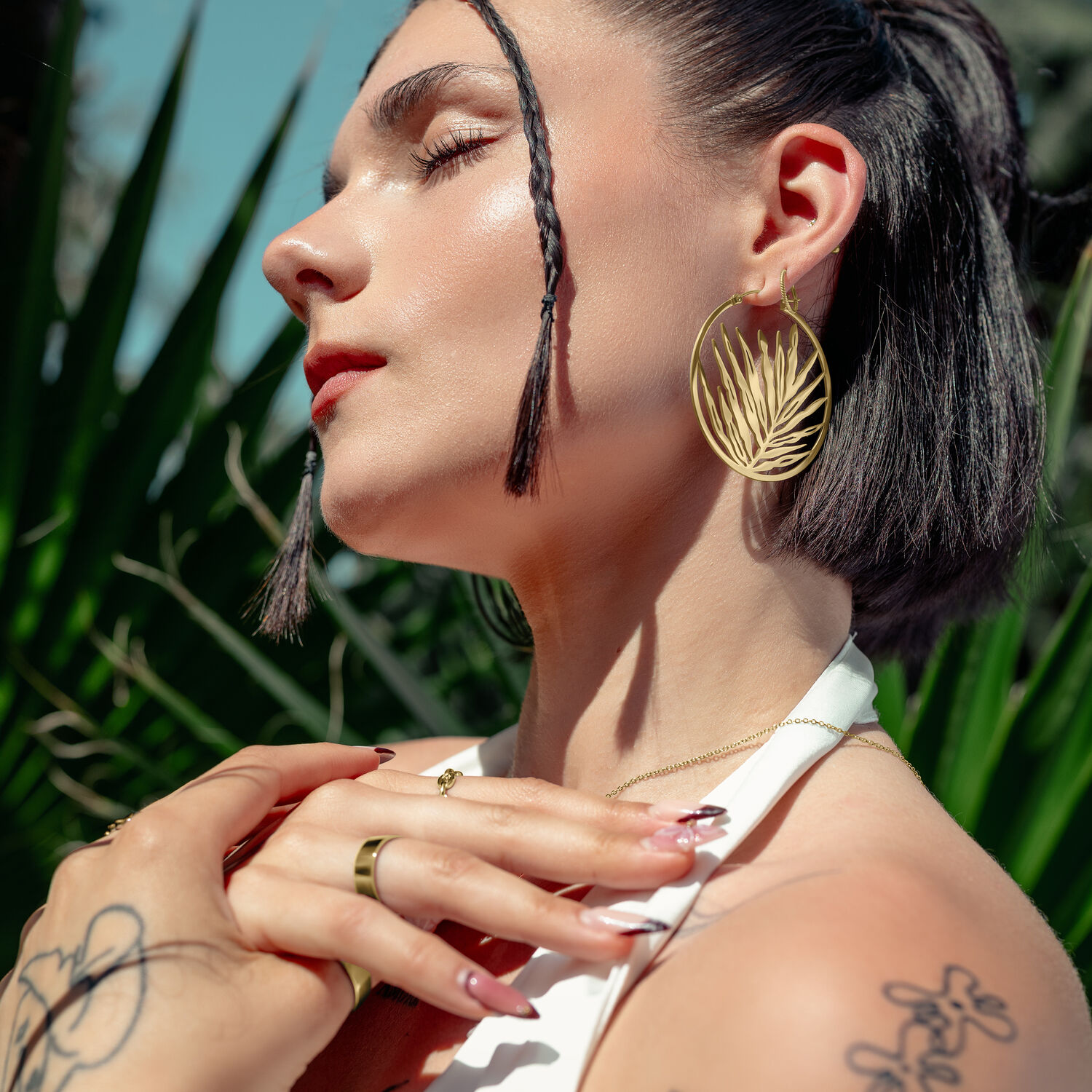 Goldene Noble Leaf Hoops am Ohr Frau mit goldenen Ohrringen in Blattform, draußen vor grünen Pflanzen