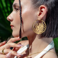 Goldene Noble Leaf Hoops Ohrringe im Sommerlook Frau mit goldenen Noble Leaf Hoops Ohrringen, im Profil vor grünen Blättern