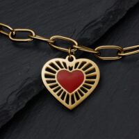 Goldenes Sacred Heart Armband mit rotem Herz Armband aus goldfarbenem Metall mit herzförmigem Anhänger und rotem Herz auf Schieferhintergrund