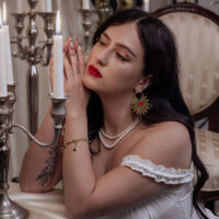 Goldene Ohrringe Sacred Heart im Kerzenlicht Frau mit goldenen Sacred Heart Ohrringen, rote Lippen, Kerzen im Hintergrund