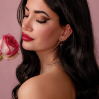 Pitkähiuksinen tumma nainen pitää vaaleanpunaista ruusua ja käyttää hopeista korvakorua punaisella kristallilla vaaleanpunaisella taustalla