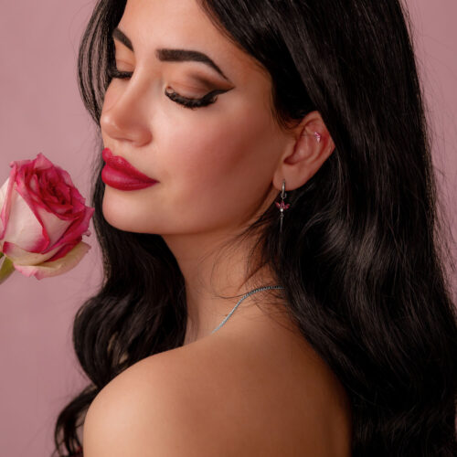 Pitkähiuksinen tumma nainen pitää vaaleanpunaista ruusua ja käyttää hopeista korvakorua punaisella kristallilla vaaleanpunaisella taustalla