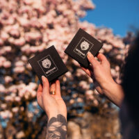 Kaksi mustaa laatikkoa Wildcat-logolla, käsissä pidettynä, kukkivan puun edessä