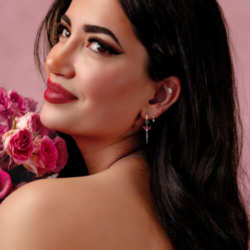 Pitkähiuksinen nainen käyttää hopeisia korvakoruja punaisilla kristalleilla ja pitää vaaleanpunaisia ruusuja vaaleanpunaista taustaa vasten.