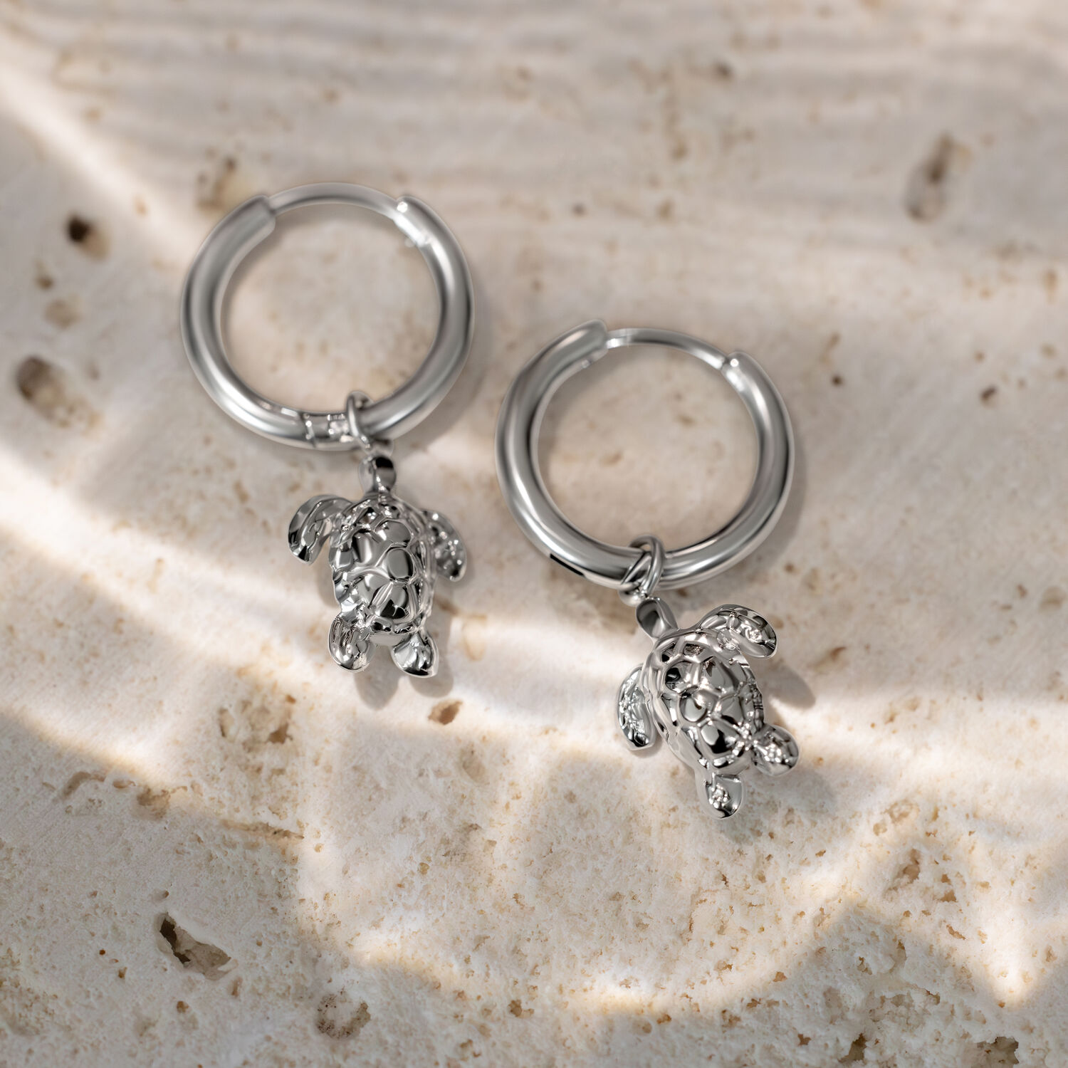 Kaksi hopeista korvarengasta pienillä kilpikonna-riipuksilla vaalealla kivialustalla