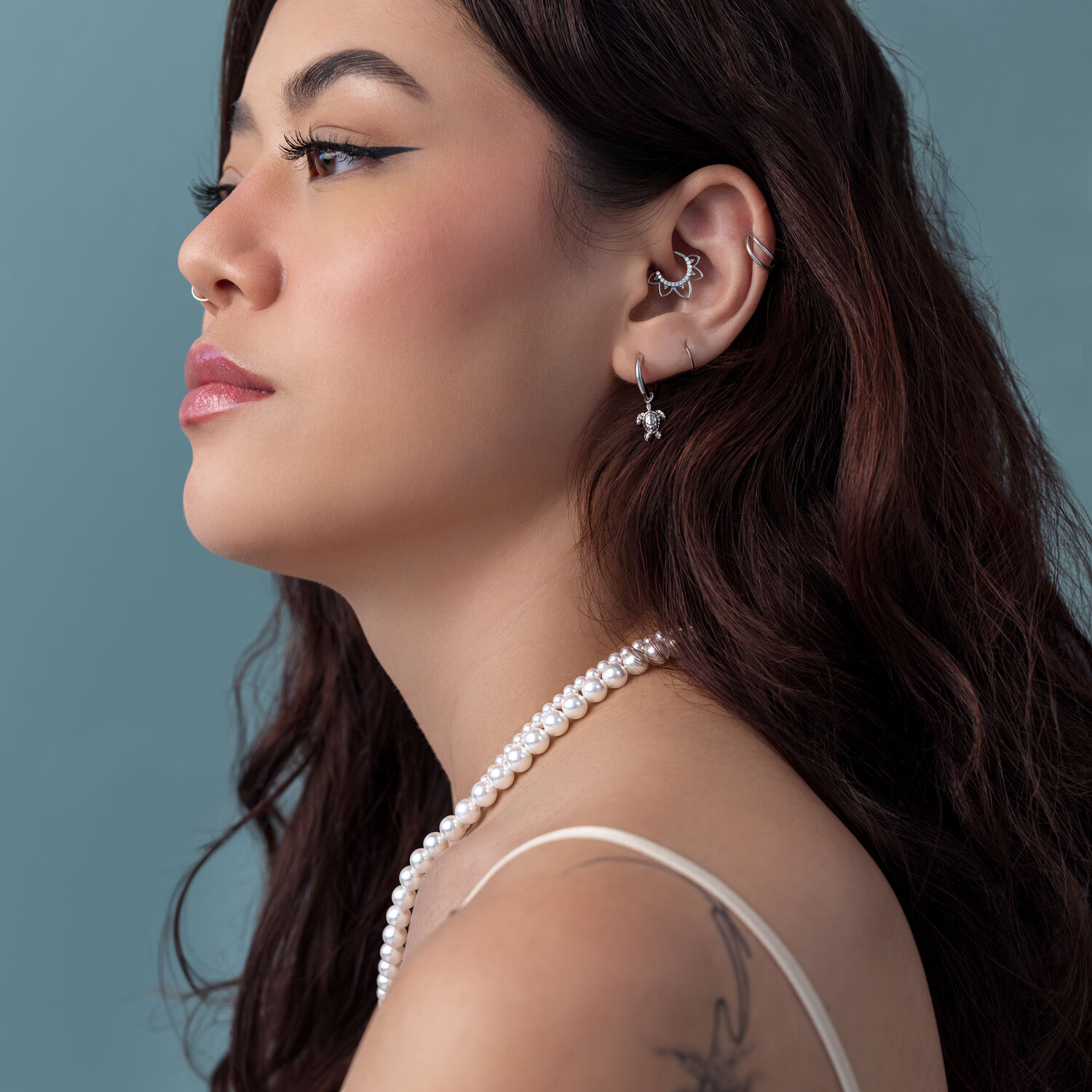 Nainen, jolla on pitkät ruskeat hiukset, hopeiset Turtle-korvakorut, helmiketju, sininen tausta