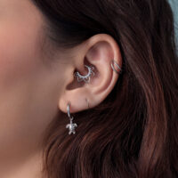 Lähikuva korvasta, jossa on hopeiset kilpikonna-korvakorut ja muita lävistyksiä, taustalla ruskeat hiukset