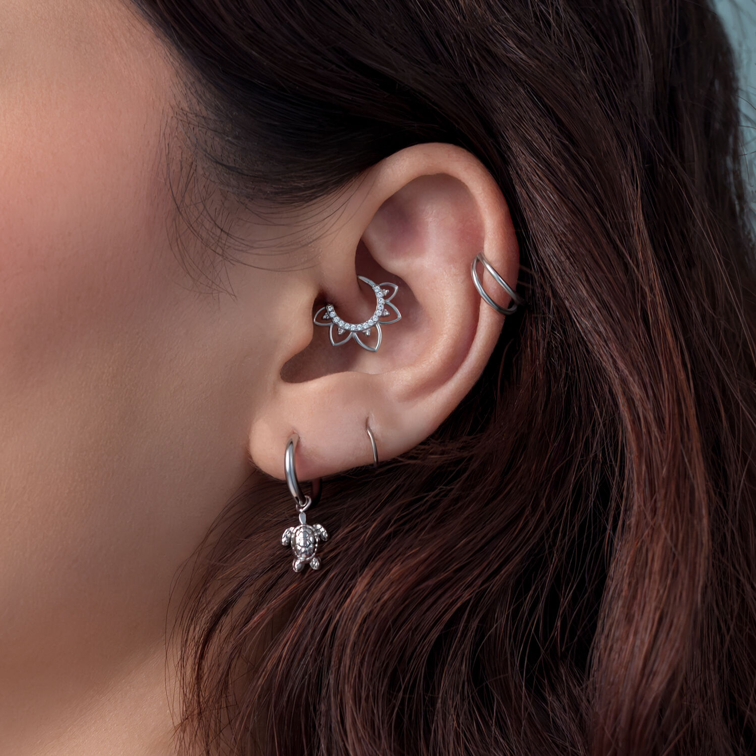 Lähikuva korvasta, jossa on hopeiset kilpikonna-korvakorut ja muita lävistyksiä, taustalla ruskeat hiukset