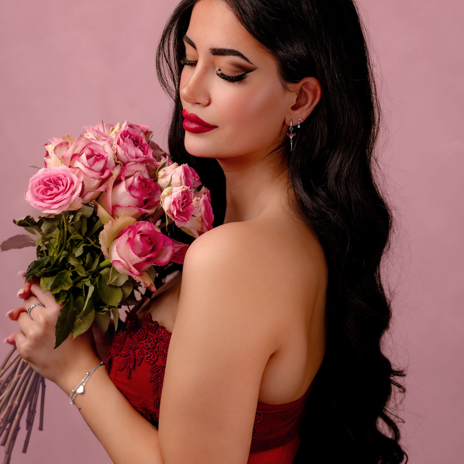 Pitkähiuksinen, tumma nainen pitää kimppua vaaleanpunaisia ruusuja vaaleanpunaista taustaa vasten.