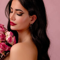 Nainen, jolla on pitkät tummat hiukset ja hopeiset korvakorut punaisilla kristalleilla, pitää vaaleanpunaisia kukkia vaaleanpunaista taustaa vasten