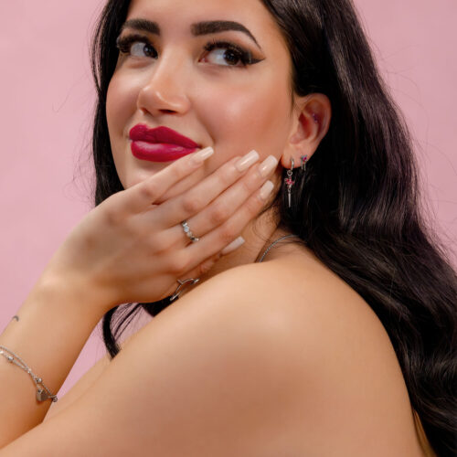 Pitkähiuksinen tumma nainen, joka käyttää hopeisia koruja punaisilla kristalleilla vaaleanpunaista taustaa vasten