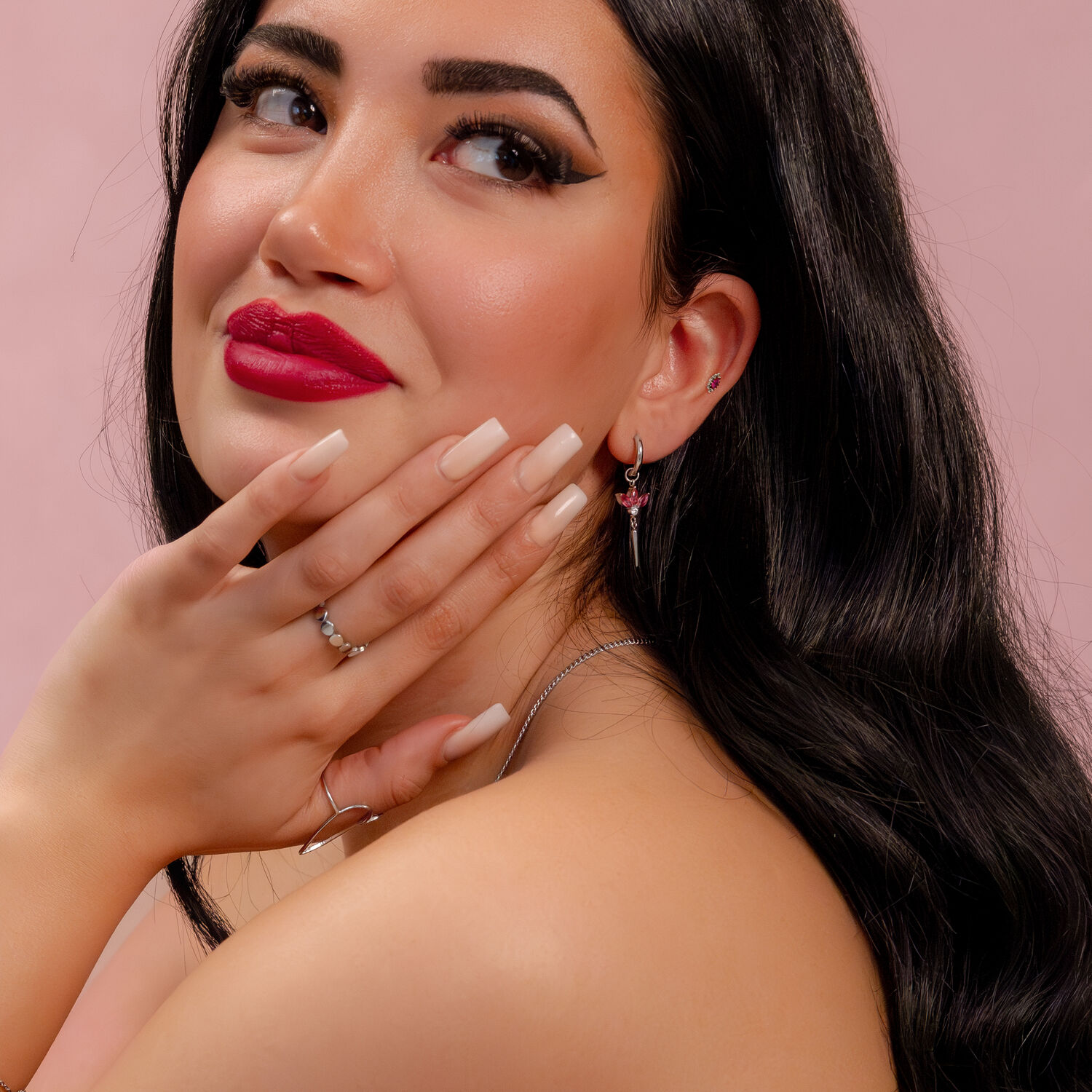 Nainen, jolla on pitkät mustat hiukset, punaiset huulet ja hopeinen korvakoru punaisella kristallilla vaaleanpunaista taustaa vasten