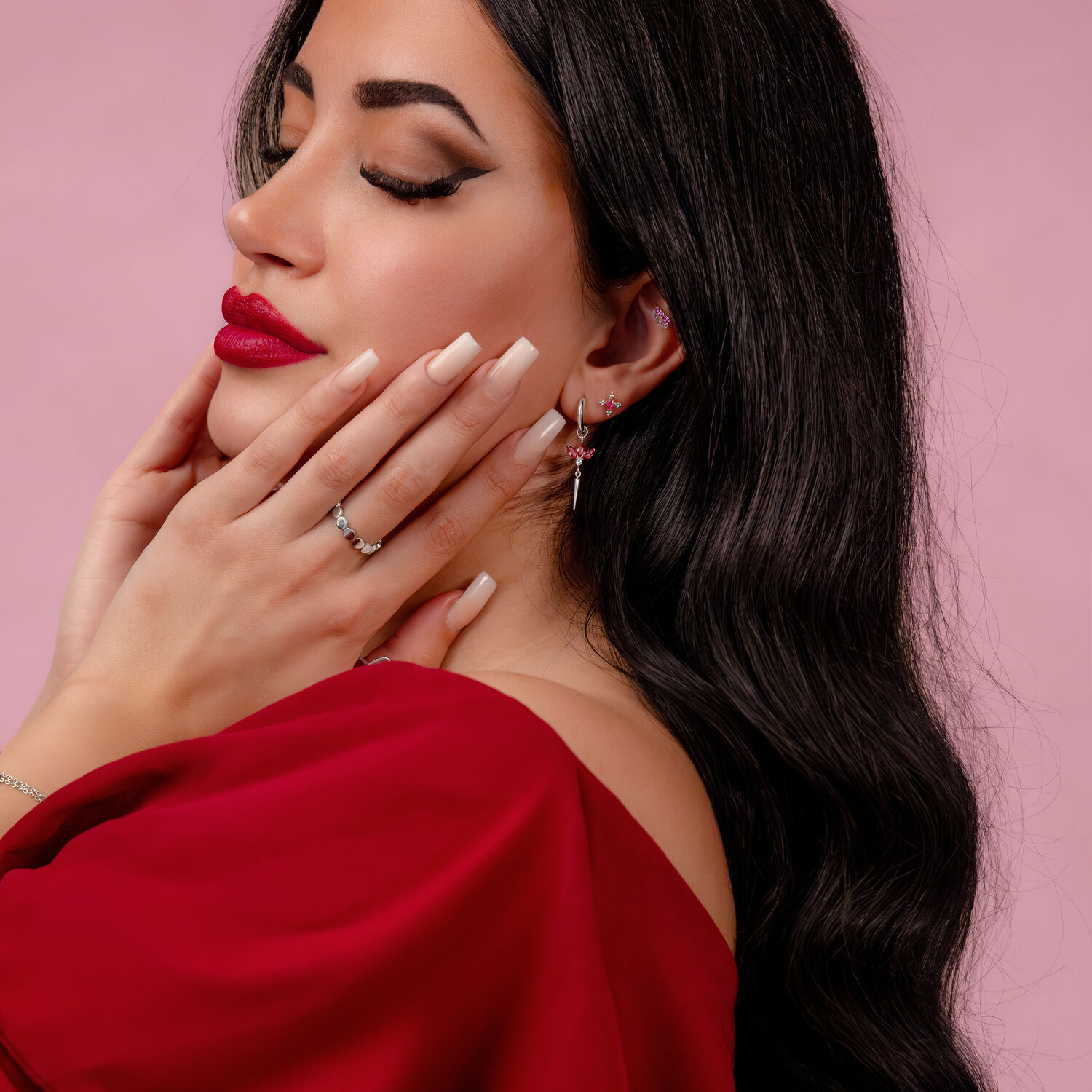 Nainen, jolla on punaiset huulet ja pitkät, suorat hiukset, käyttää hopeista korvakorua vaaleanpunaisella taustalla