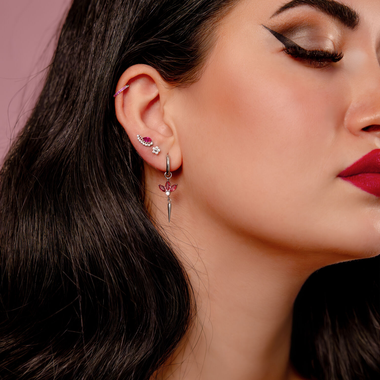 Lähikuva korvasta, jossa on useita lävistyksiä, mukaan lukien hopeinen clicker punaisilla kristalleilla, vaaleanpunaisella taustalla.