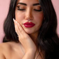 Nainen mustilla hiuksilla ja hopeisella nenäkorulla, jossa on punainen kristalli, vaaleanpunaista taustaa vasten