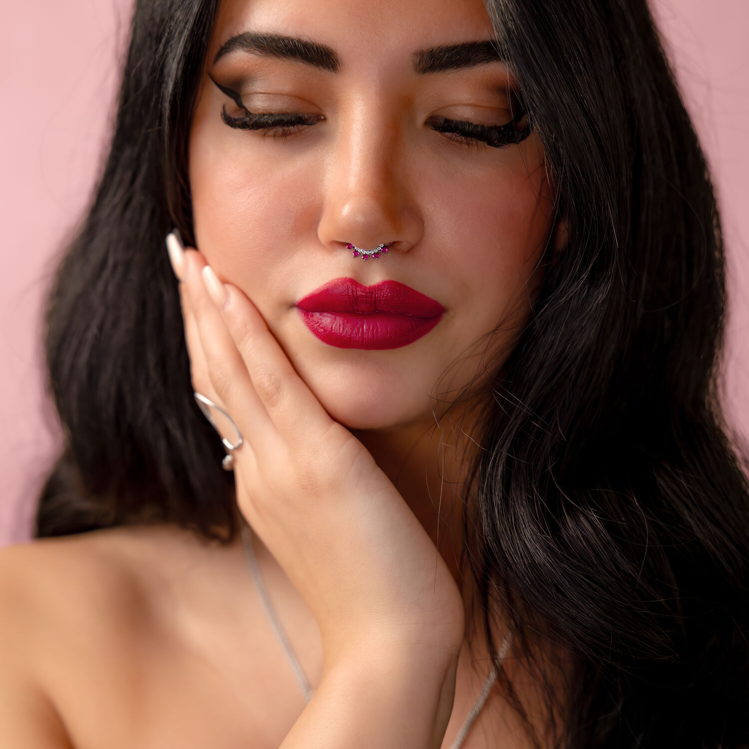 Nainen mustilla hiuksilla ja hopeisella nenäkorulla, jossa on punainen kristalli, vaaleanpunaista taustaa vasten