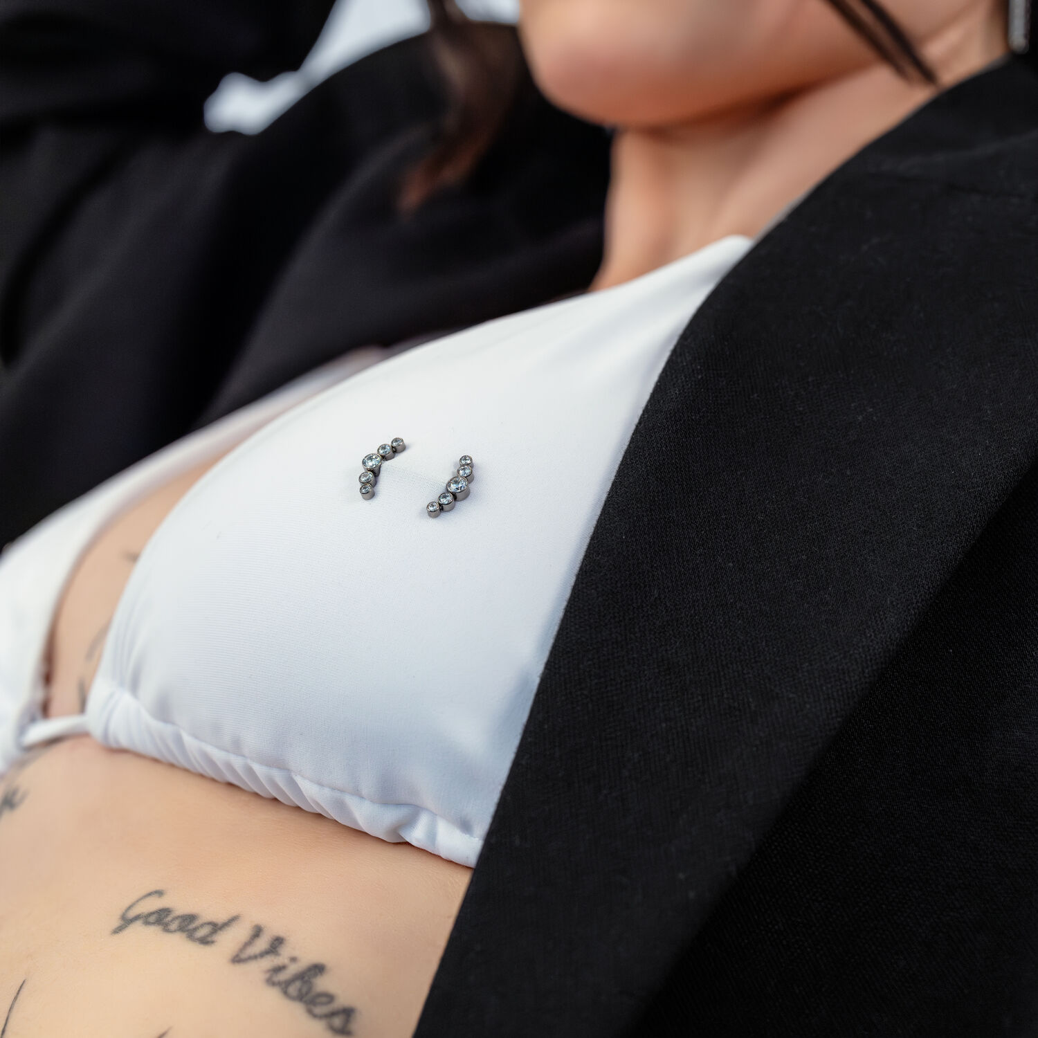 Lähikuva valkoisesta bikinien yläosasta Penta Crystal Barbell -lävistyksellä ja tatuoinnit taustalla.